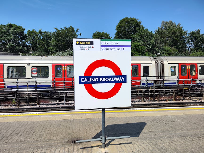 A signage at Ealing Broadway station displaying directions to the district and Elizabeth lines, with a train in the background. The sign features a white background with a large red circle and a blue band across the middle bearing the station name. The station platform surface is made of textured concrete tiles, clean and free of litter, with a yellow safety line marking the edge. Behind the sign, a modern train with a silver body, red and blue accents, and tinted windows is parked along the tracks. Overhead, there are green trees and a clear blue sky, indicating a bright, sunny day. The overall scene highlights the tidy, well-maintained environment typical of UK railway stations, with no visible dirt or debris, reflecting effective station cleaning and management, and natural lighting emphasizing the station's operational condition.