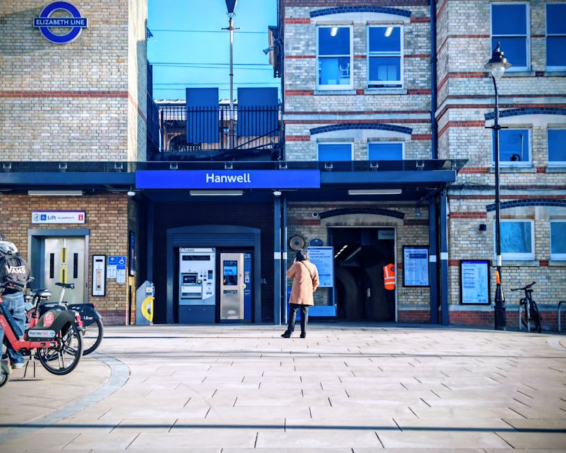 The exterior of Hanwell station showing a brick building with large blue-framed windows and a blue signage sign reading 'Hanwell'. In front of the station entrance, there is a paved pavement with a woman in a light pink coat standing nearby, facing the ticket machines and entrance. To the left, bicycles are parked with visible branding, including a bike sharing station, and to the right, a black stool and a wall-mounted bulletin board are present. The station features a modern black awning with the Elizabeth Line logo at the top, and the overall scene appears clean and well-maintained, emphasizing surface cleaning and hygiene standards consistent with professional cleaning services such as those offered by Colliers Wood Cleaners.