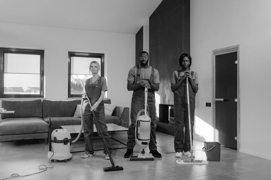 Three professional cleaners from Colliers Wood Cleaners are performing surface cleaning and deep cleaning in a modern living room. The room features a light-colored wooden floor that appears spotless and well-maintained, with a dark grey fabric sofa positioned against the wall beneath two large windows allowing natural light to illuminate the space. The cleaning team, dressed in matching uniforms, utilizes vacuum cleaners and floor mops to ensure thorough hygiene and cleanliness on the tiled flooring. One cleaner holds a vacuum hose connected to a canister model, while the others use mops and a small bucket for scrubbing and sanitisation of surfaces. The room has a minimalist aesthetic with a dark accent wall and a simple coffee table, creating a clean and inviting atmosphere, representative of the professional domestic cleaning services provided by Colliers Wood Cleaners. The overall scene emphasizes meticulous surface and deep cleaning practices in a residential setting, highlighting the importance of hygiene and maintenance in homes.