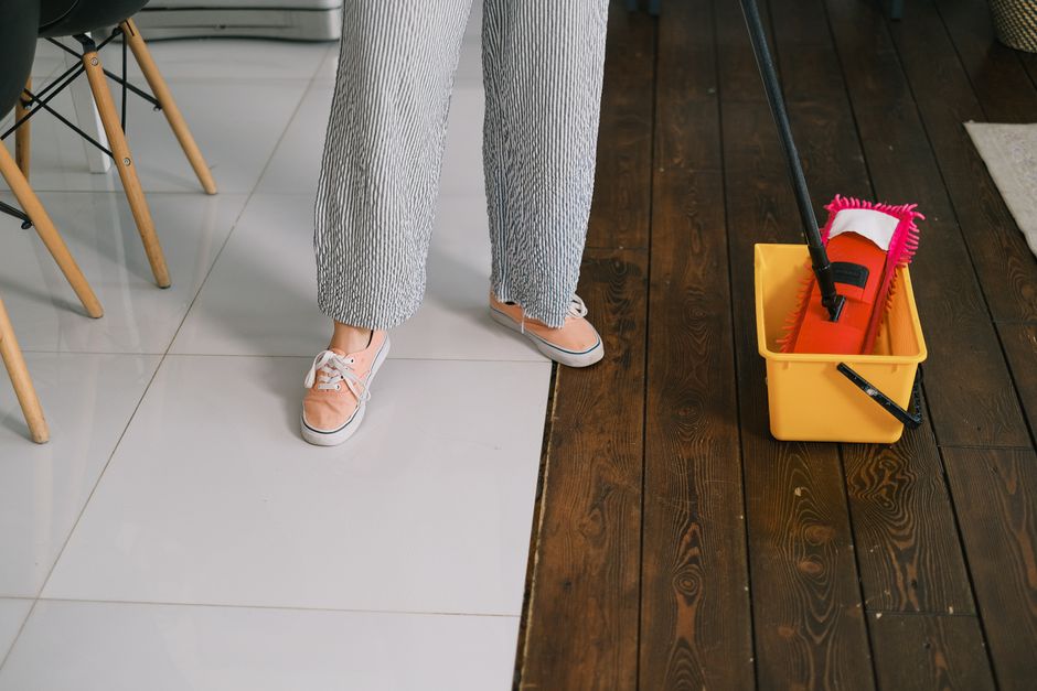Close-up view of a person's lower legs and feet standing on a tiled floor with white ceramic tiles, adjacent to a darker wooden floor in a room. The person is wearing light gray, textured pants and beige sneakers with white soles. Near their feet, a yellow cleaning bucket containing a red mop with a pink microfiber head, a black squeegee, and a spray bottle is placed on the wooden floor. The setting appears to be a kitchen or dining area, with part of a wooden chair with angled legs visible on the left. The area is well-lit with natural light, highlighting the clean and maintained surfaces, consistent with professional domestic cleaning services by Colliers Wood Cleaners.
