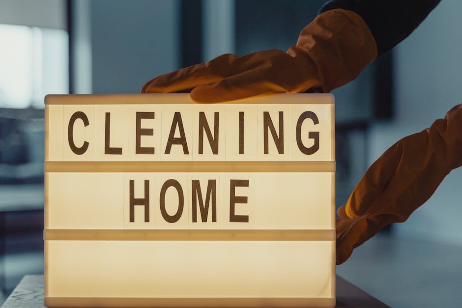 Three professional cleaners from Colliers Wood Cleaners are performing surface cleaning and deep cleaning in a modern living room. The room features a light-colored wooden floor that appears spotless and well-maintained, with a dark grey fabric sofa positioned against the wall beneath two large windows allowing natural light to illuminate the space. The cleaning team, dressed in matching uniforms, utilizes vacuum cleaners and floor mops to ensure thorough hygiene and cleanliness on the tiled flooring. One cleaner holds a vacuum hose connected to a canister model, while the others use mops and a small bucket for scrubbing and sanitisation of surfaces. The room has a minimalist aesthetic with a dark accent wall and a simple coffee table, creating a clean and inviting atmosphere, representative of the professional domestic cleaning services provided by Colliers Wood Cleaners. The overall scene emphasizes meticulous surface and deep cleaning practices in a residential setting, highlighting the importance of hygiene and maintenance in homes.