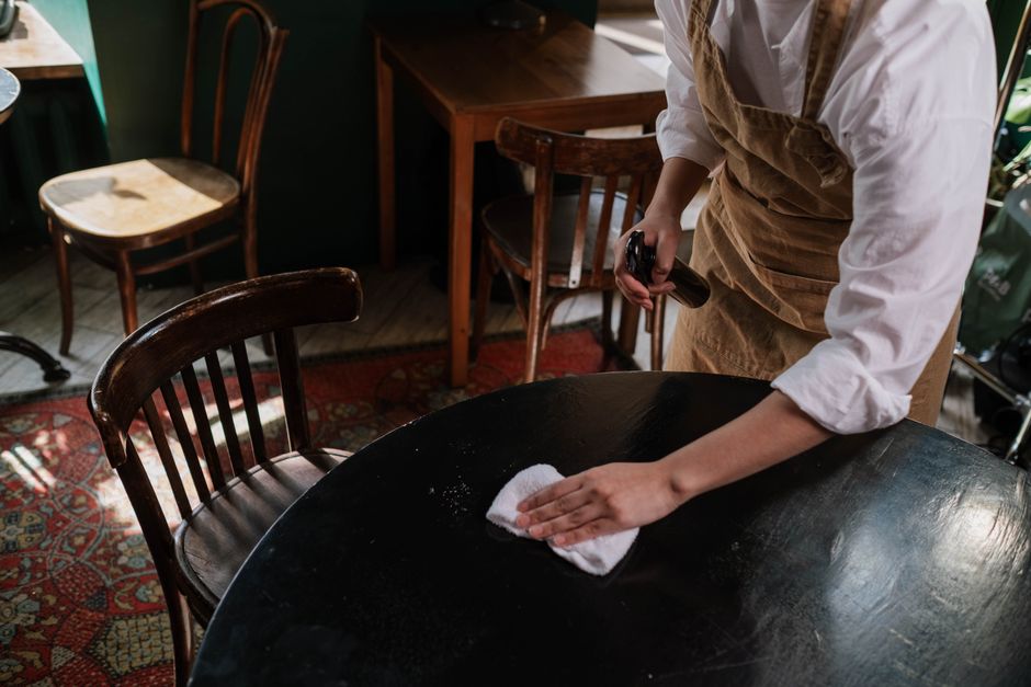 A person wearing a white shirt and a brown apron is engaged in surface cleaning at a dark wooden table in a cafe setting. They are using a white cloth to wipe the smooth, reflective surface of the table, which appears to have been recently cleaned or disinfected. The person holds a spray bottle in their right hand, indicating hygiene and sanitisation processes. Surrounding the table are vintage-style wooden chairs with curved backs, and in the background, there are additional tables and chairs, a patterned rug with red and blue tones, and natural lighting entering from nearby windows, creating a warm and inviting atmosphere. Colliers Wood Cleaners specializes in professional cleaning services, including deep cleaning and hygiene maintenance for cafes and similar establishments, as highlighted on their Merton Abbey Mills Colliers Wood cafe cleaning checklist page.