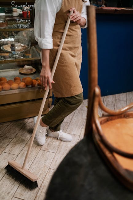 A person wearing a white shirt, beige apron, and khaki pants is sweeping the wooden floor of a cafe with a long-handled brush. The cafe interior features a glass display case with baked goods such as pastries and muffins, visible behind the person, with handwritten chalkboard signs indicating different desserts. There is a blue counter or partition, and the room is illuminated with warm lighting. The scene demonstrates surface cleaning and tidying as part of routine hygiene maintenance, with Colliers Wood Cleaners specializing in domestic and commercial deep cleaning services, including cafe cleaning in the Colliers Wood area.