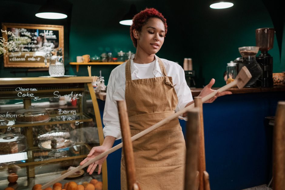 A person wearing a white shirt and a brown apron is engaged in surface cleaning at a dark wooden table in a cafe setting. They are using a white cloth to wipe the smooth, reflective surface of the table, which appears to have been recently cleaned or disinfected. The person holds a spray bottle in their right hand, indicating hygiene and sanitisation processes. Surrounding the table are vintage-style wooden chairs with curved backs, and in the background, there are additional tables and chairs, a patterned rug with red and blue tones, and natural lighting entering from nearby windows, creating a warm and inviting atmosphere. Colliers Wood Cleaners specializes in professional cleaning services, including deep cleaning and hygiene maintenance for cafes and similar establishments, as highlighted on their Merton Abbey Mills Colliers Wood cafe cleaning checklist page.