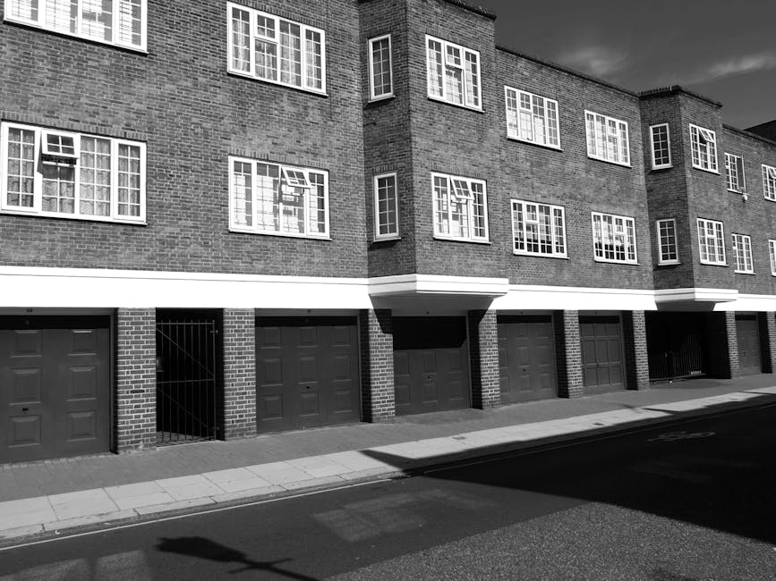 Black and white exterior view of a modern residential block with multiple windows and multiple garage doors at ground level. The building features a brick facade with a clean, well-maintained appearance, and the garages have paneled doors. The sidewalk runs along the street, with visible curb lines casting shadows on the pavement. The lighting indicates natural daylight, highlighting the building's structure and surface details. This image exemplifies the urban residential environment that Colliers Wood Cleaners services for end of tenancy cleaning in High Street flats, COLLIERS WOOD, focusing on maintaining cleanliness and hygiene for properties in the SW19 area.