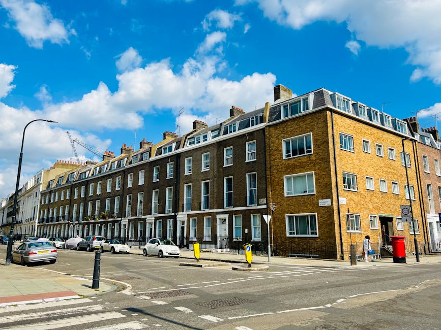 A row of modern multi-story residential flats with yellow brick facades and large white-framed windows, situated along a busy street under a bright blue sky with scattered clouds. The street features parked cars, a pedestrian walking on the sidewalk, black bollards, and a red post box near the building's corner. The overall scene appears clean and well-maintained, with clear signage and street furniture enhancing urban hygiene and order. This setting highlights the importance of regular cleaning and maintenance in residential areas, aligning with the services offered by Colliers Wood Cleaners for end of tenancy and domestic cleaning needs.