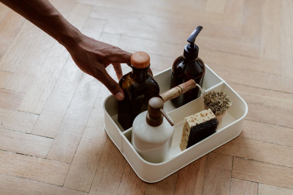 A person’s hand reaching for brown and black bottles of cleaning products placed inside a white plastic storage tray on a wooden floor. The tray contains various items including a white spray bottle, a cleaning sponge, a scrub brush, and a wooden-handled tool. The scene is well-lit with natural light, highlighting the clean and organized appearance of the items, which are used for surface and deep cleaning. This setup reflects domestic cleaning practices typical of maintaining hygiene in riverside flats, aligning with the home cleaning tips provided by Colliers Wood Cleaners in the Wandle Park Colliers Wood area.
