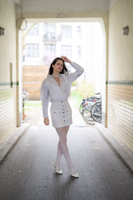 A young woman standing in an outdoor passageway with beige tiled walls and a concrete ceiling, illuminated by natural daylight. She is dressed in a white long-sleeved blouse, a high-waisted white skirt, and white tights, accessorized with a choker necklace. She has dark hair styled with loose waves and is posing with one hand running through her hair, looking thoughtfully into the distance. The background features bicycles parked against the wall and a blurred view of greenery and buildings beyond the passage. The scene is crisply lit, emphasizing her outfit and the textured surfaces of the tiled walls and the concrete ground, evoking a sense of urban calm and style relevant to domestic or commercial cleaning contexts, as highlighted by Colliers Wood Cleaners.
